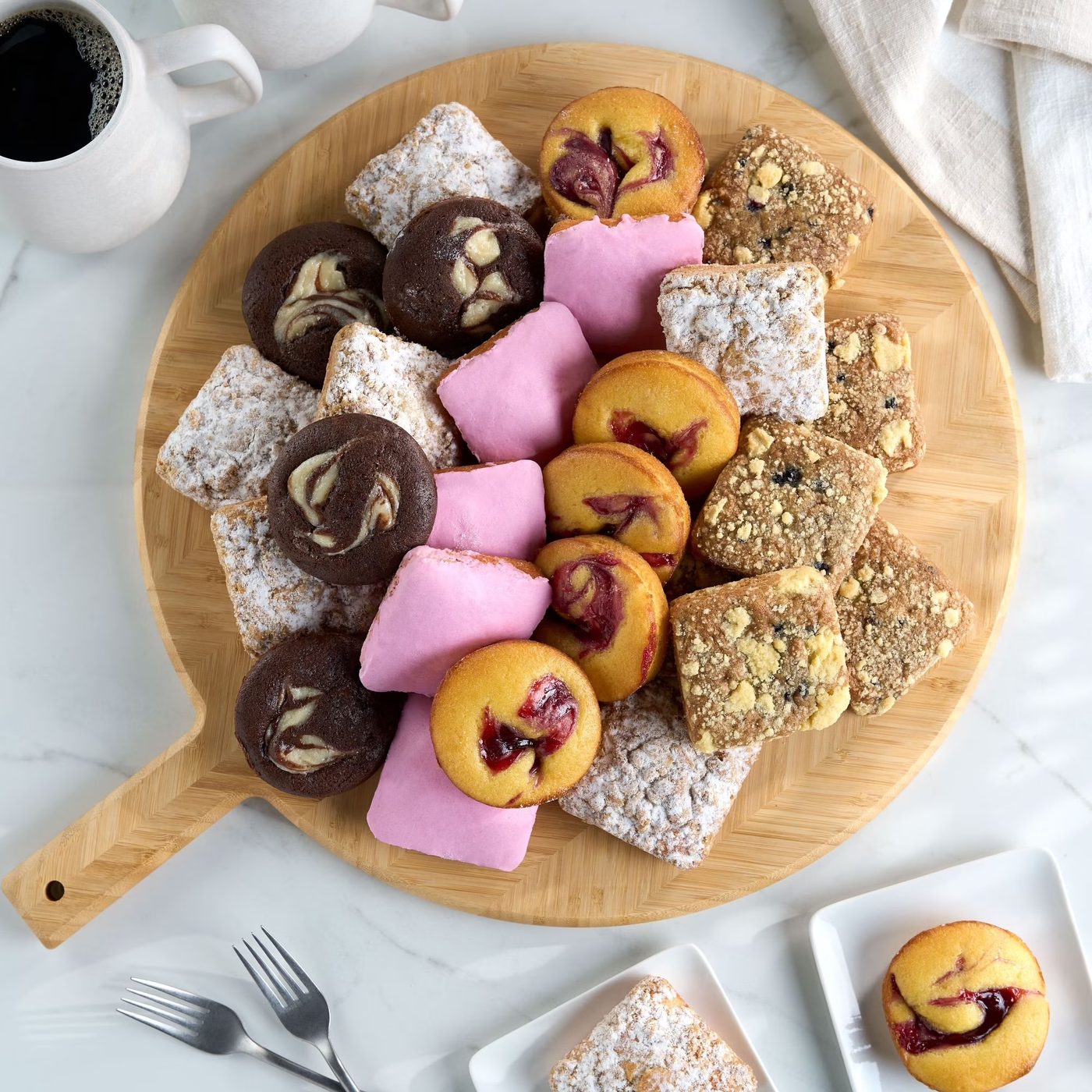 Round wooden board with mini chocolate cakes, fruit-filled pastries, powdered squares, and crumb cake bites
